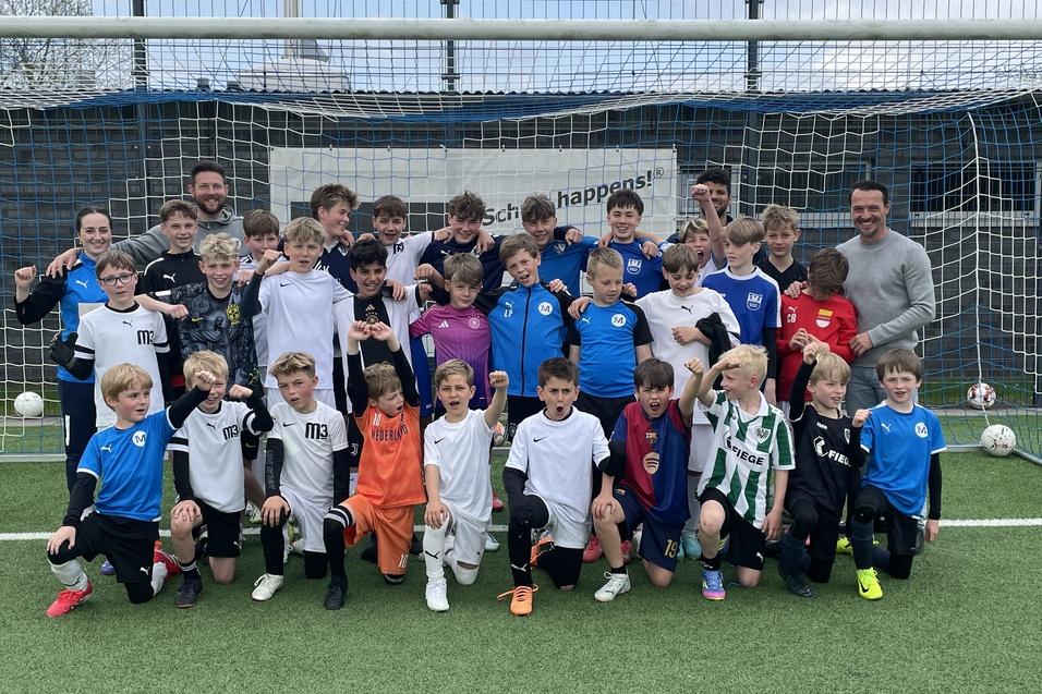 Gruppe von Kindern in Fu&szlig;balltrikots posiert l&auml;chelnd vor einem Fu&szlig;balltor auf einem Kunstrasenfeld.