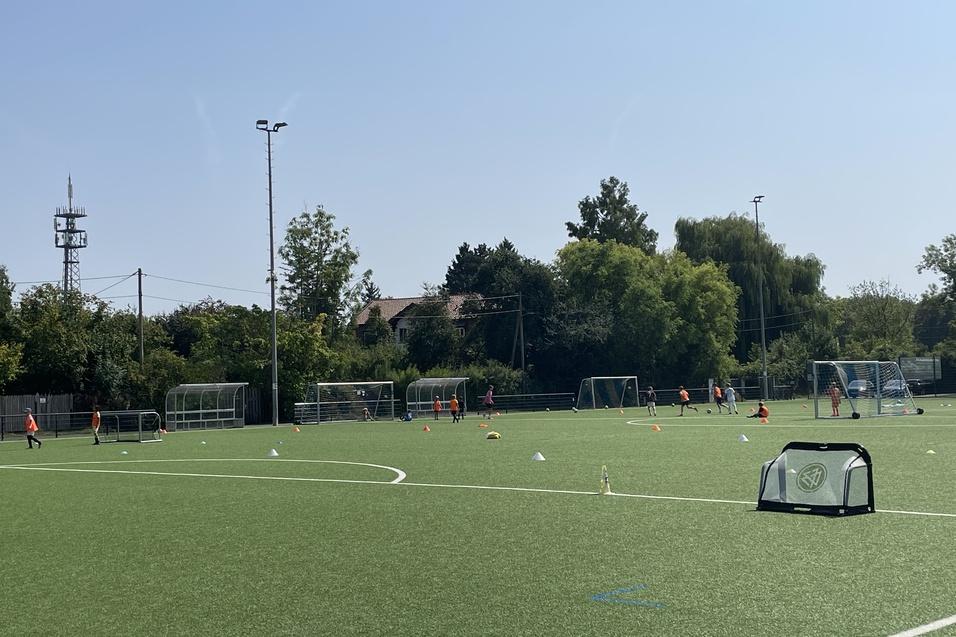 Fu&szlig;ballfelder mit Trainingszubeh&ouml;r, Spielern in oranger Kleidung und B&auml;umen unter klarem Himmel.