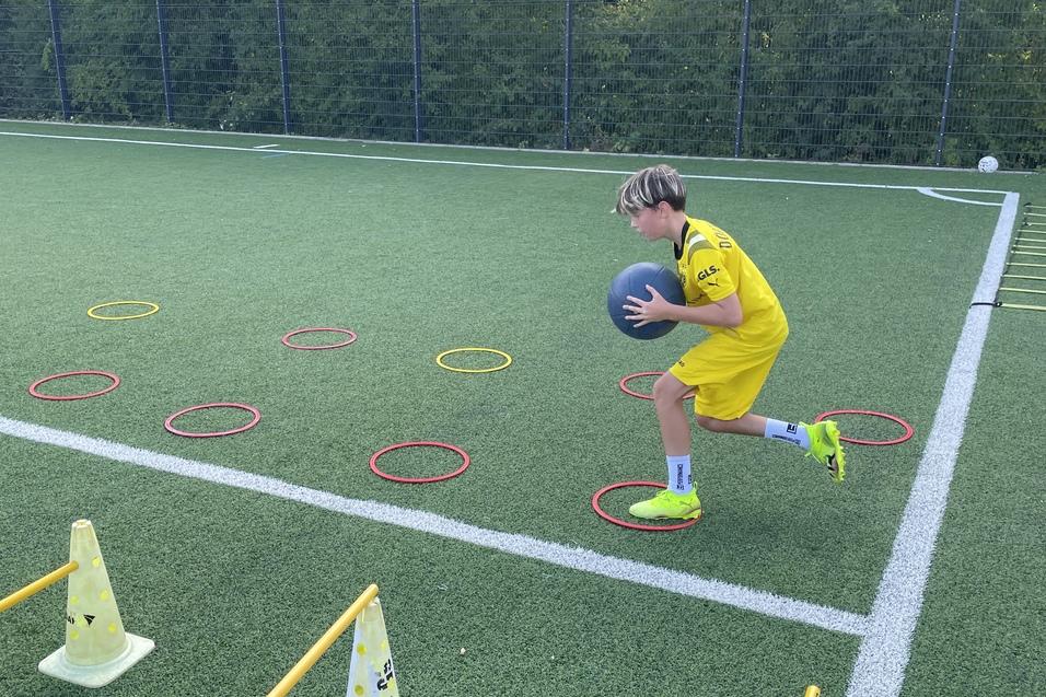 Junge in gelber Sportkleidung l&auml;uft auf Kunstrasen mit einem Medizinball zwischen H&uuml;tchen und Ringen.