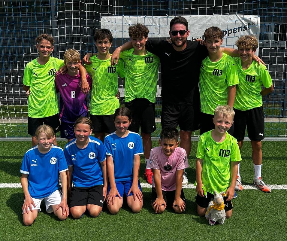 Gruppe von zehn Jugendlichen in Fu&szlig;balltrikots, posierend vor einem Tor auf einem Rasenplatz.